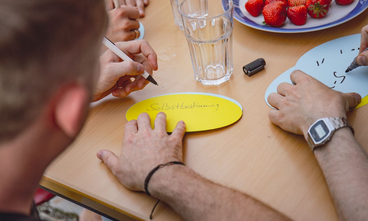 Personen schreiben auf bunte Papierkarten, wobei eine Karte mit dem Wort 'Selbstbestimmung' beschrieben wird. Auf dem Tisch stehen Gläser und ein Teller mit Erdbeeren.
