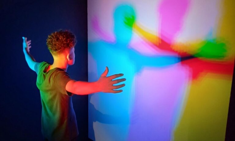 Eine Person mit ausgestreckten Armen steht vor einer Wand, auf der durch bunte Lichter farbige Schatten in Blau, Rot und Grün erzeugt werden.