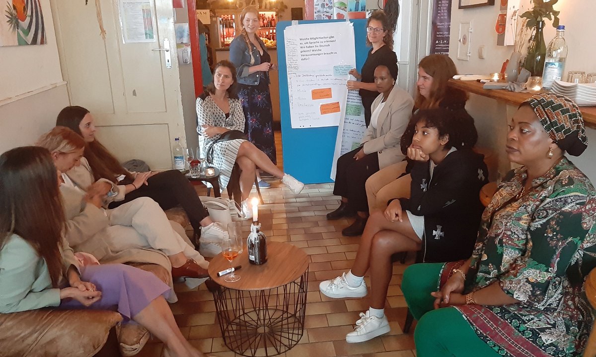 Eine Gruppe von Frauen sitzt in einem gemütlichen Raum und diskutiert, während eine Teilnehmerin eine Präsentation vor einem Flipchart hält.