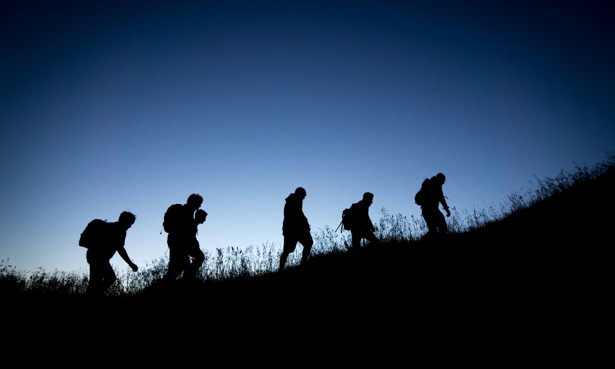 Silhouetten von Menschen mit Rucksäcken, die bei Dämmerung einen Hügel hinaufwandern, vor einem klaren, dunkelblauen Himmel.