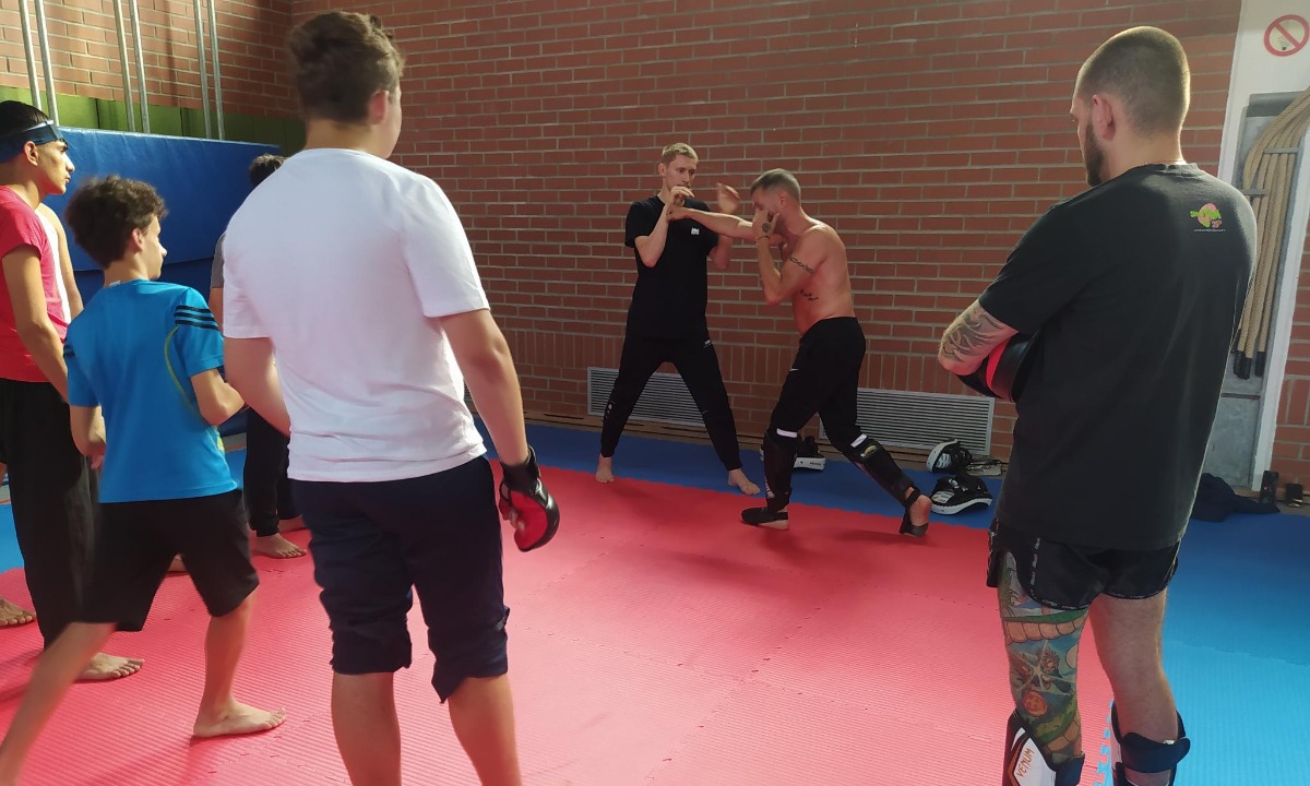 Eine Gruppe von Personen beobachtet ein Kampfkunst-Training, bei dem zwei Teilnehmer in einer Kampfszene auf einer roten Matte in einer Halle mit Backsteinwänden zu sehen sind.