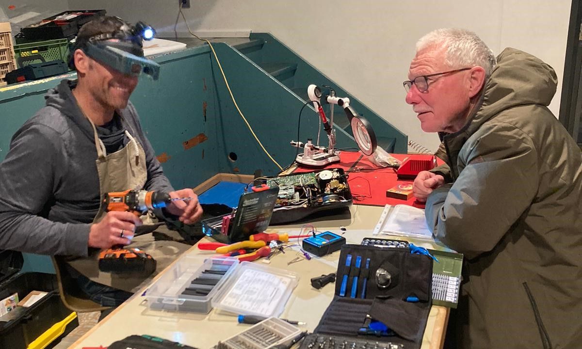 Zwei Männer sitzen an einem Arbeitstisch voller Werkzeuge und Elektronik. Einer trägt eine vergrößernde Kopflupe und hält eine Bohrmaschine, während der andere interessiert zuschaut.