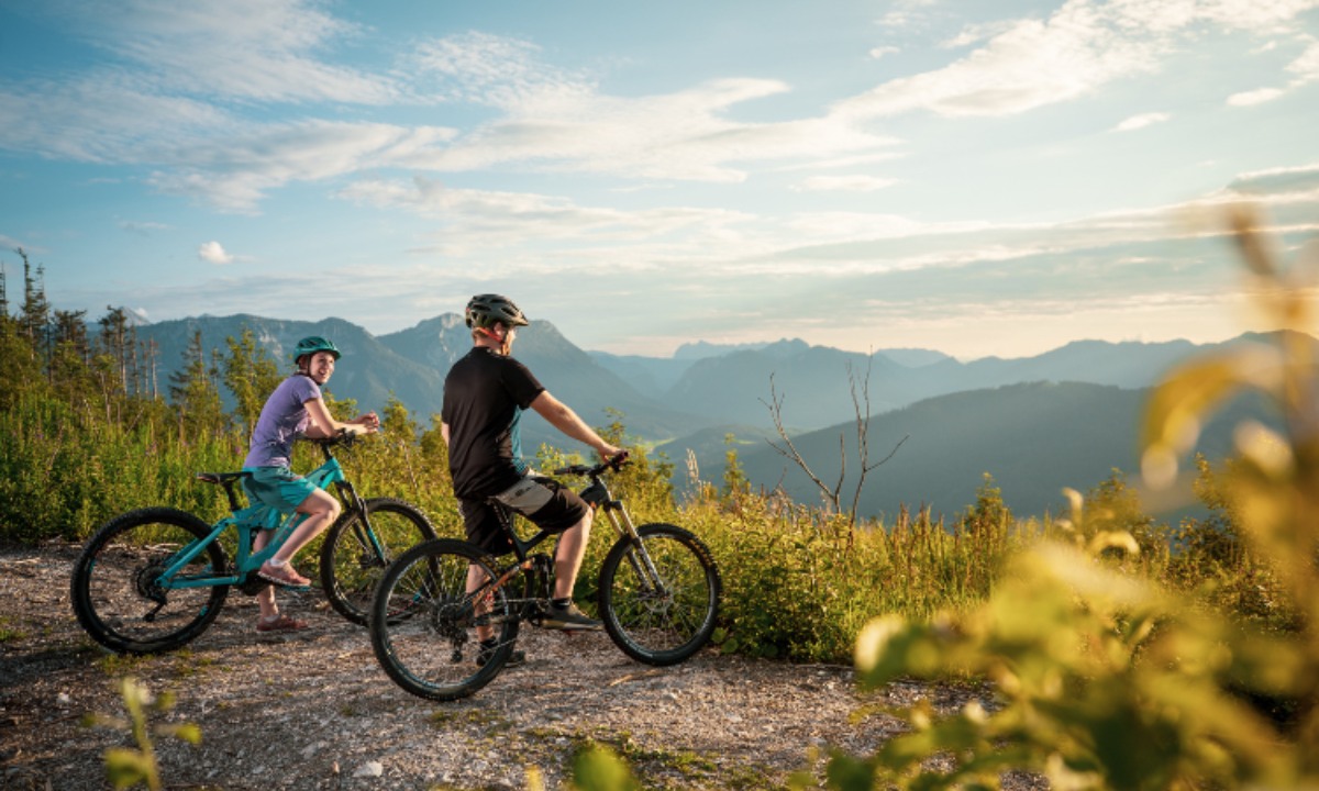 Zwei Radfahrer auf einem Bergpfad mit malerischem Blick auf eine bergige Landschaft bei Sonnenuntergang.