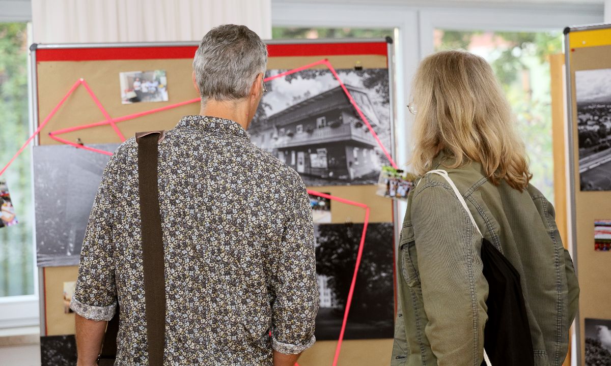 Zwei Personen betrachten eine Pinnwand mit Schwarz-Weiß-Fotografien und roten Markierungen, die möglicherweise eine Ausstellung oder Präsentation darstellt.