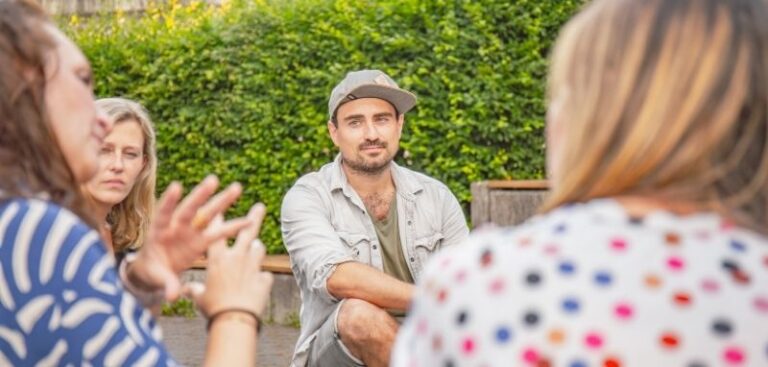 Vier Personen sitzen sich gegenüber. Sie haben bunte Kleidung an und im Fokus ist eine Person die eine Cap auf hat.