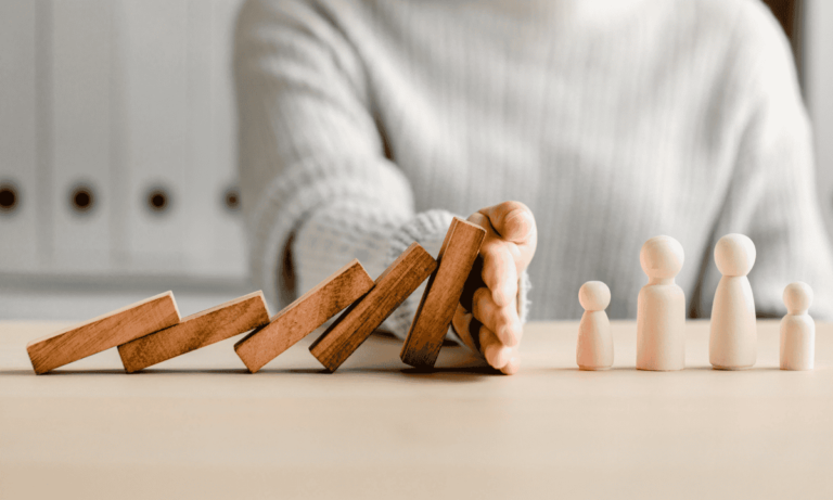 Symbolbild für Prävention - eine Reihe von Holzsteinchen fällt um, eine Hand hält sie auf, bevor sie kleine Spielfiguren umwerfen.