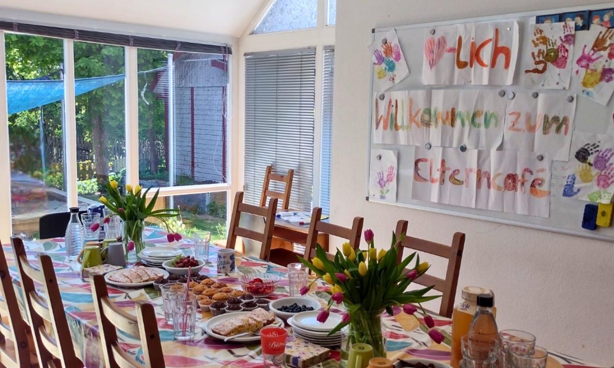 In einem Wohnraum steht ein bunt gedeckter hölzerner Tisch, an der Wand hängt ein selbst gebasteltes Plakat das besagt "Herzlich Willkommen zum Elterncafé".