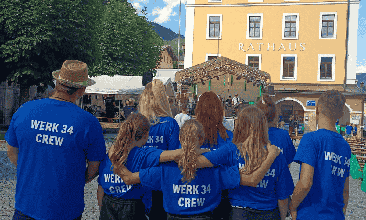 Acht Gruppenmitglieder der „WERK 34 CREW" stehen mit dem Rücken zur Kamera vor einer Bühne und lauschen der spielenden Band.